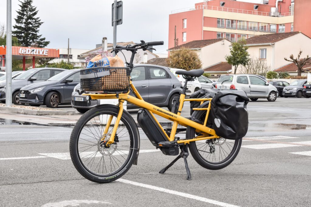 Yellow electric cargo bike carrying groceries in a front basket and side pannier bags parked in a shopping center parking lot.