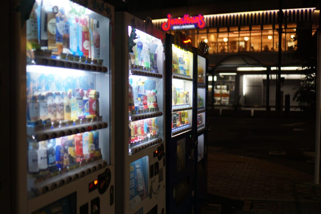 Row of illuminated vending machines stocked with drinks and snacks at night outside a shopping center.