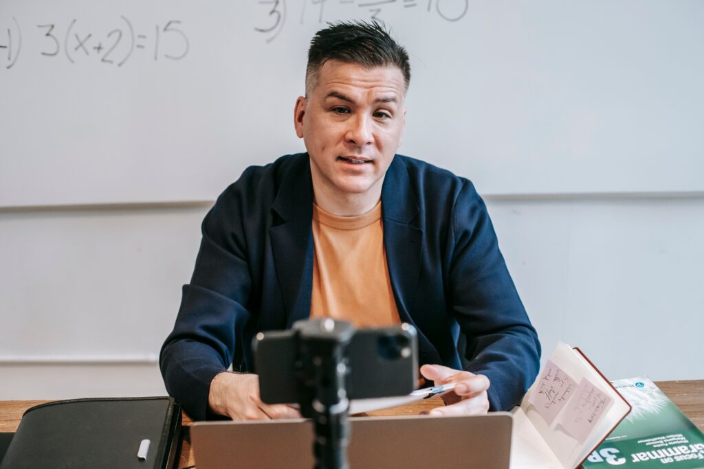 Teacher filming an online lesson with a smartphone and laptop in a classroom, illustrating remote teaching and side hustle opportunities for educators.