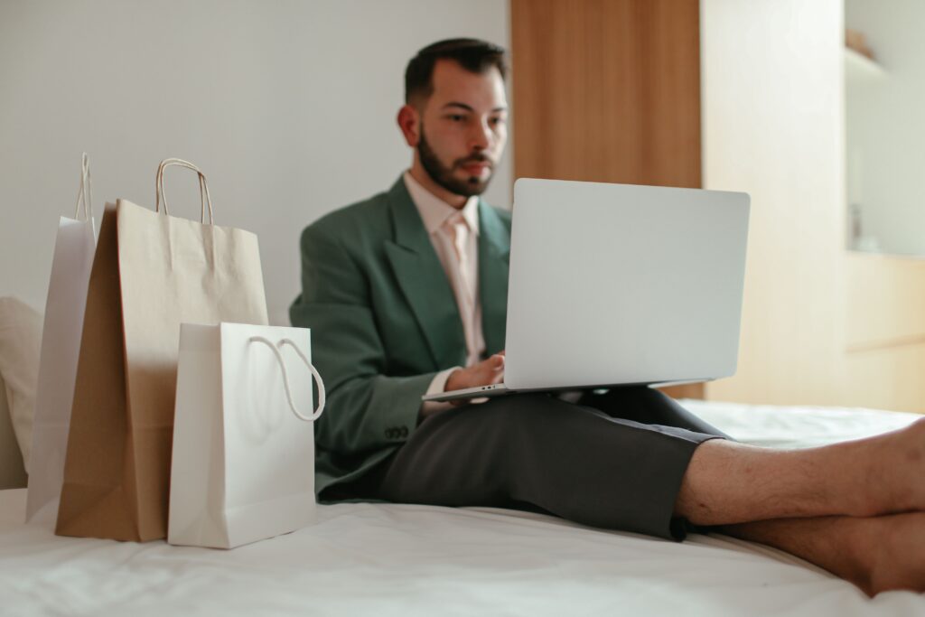 Man shopping online on a laptop while sitting on a bed with shopping bags beside him.