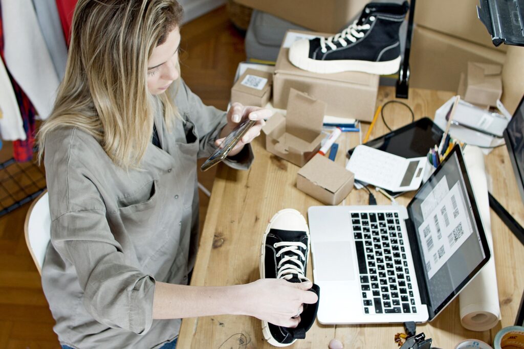 Reseller photographing a pair of sneakers for an online marketplace listing using a smartphone, with shipping boxes and a laptop on the desk.