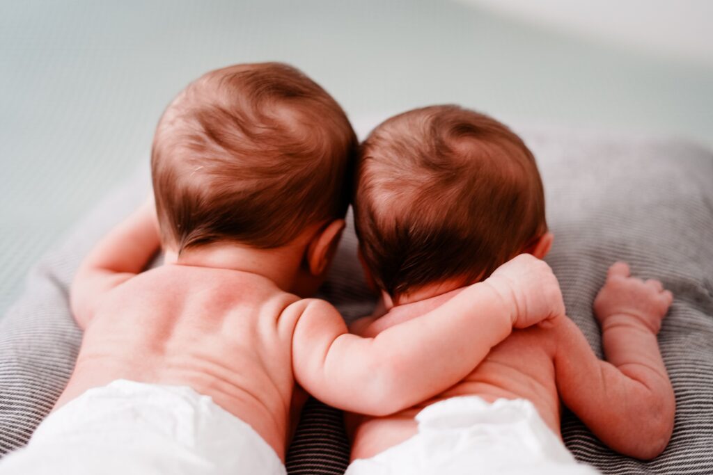 Two newborn twins lying side by side on a gray blanket, with one baby wrapping an arm around the other.