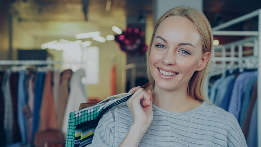 Smiling woman holding shopping bags inside a clothing store, representing retail arbitrage and buying discounted products to resell for profit.