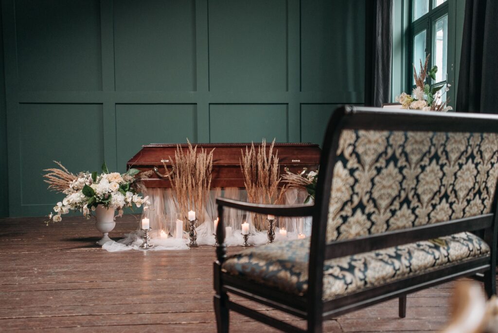 Wooden casket surrounded by flowers and candles in a funeral home with seating for mourners.