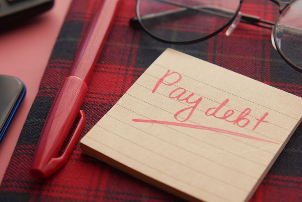 Notepad with the words "Pay Debt" written in red, alongside a pen and glasses, representing debt management and financial planning.